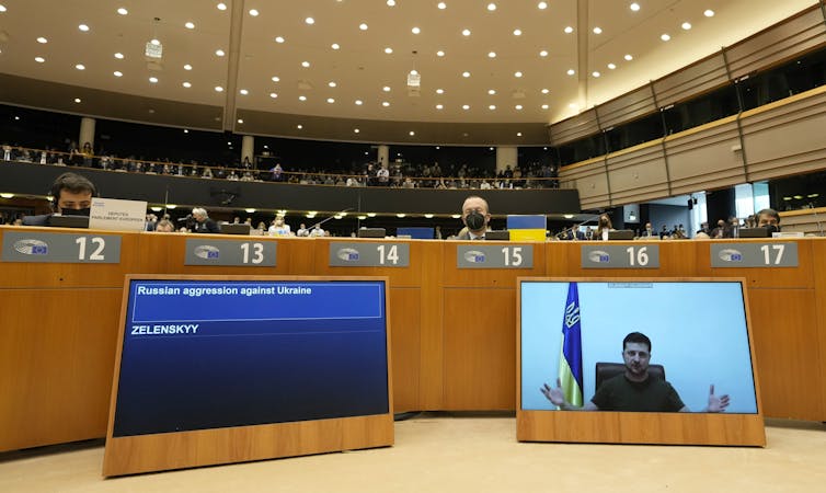 A man in a T-shirt is seen talking on a screen in a large meeting hall.
