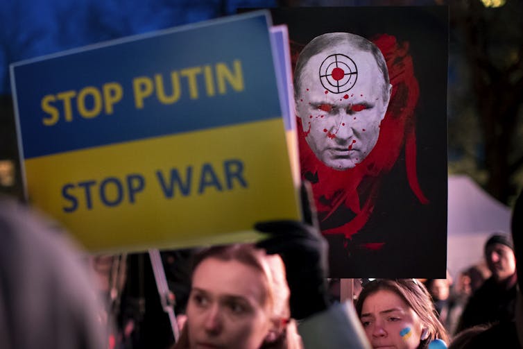 Protesters hold up a sign that reads Stop Putin, Stop War and one that portrays Putin as a demon.