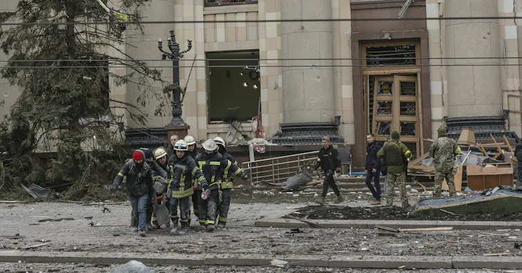 Emergency workers carry a body from a shelled building, debris surrounding them.