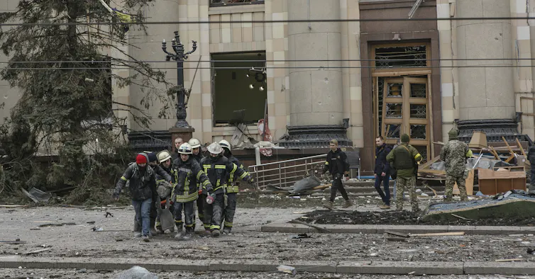 Emergency workers carry a body from a shelled building, debris surrounding them.