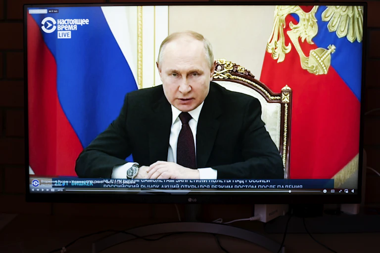 a computer screen displays a television broadcast of an older man in a dark business suit seated at a desk with a pair of flags behind him