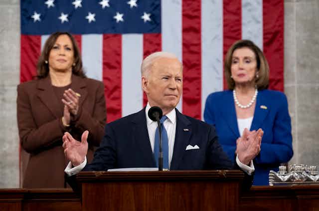 President Joe Biden stands at the podium, arms outstretched. Behind him stands Vice President Kamala Harris and House speaker Nancy Pelosi 