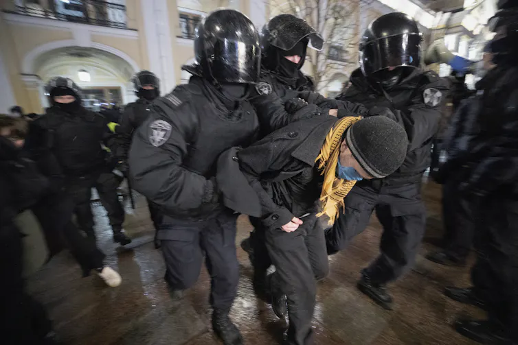 Police in heavy riot gear detaining a young person in a black hat and yellow scarf