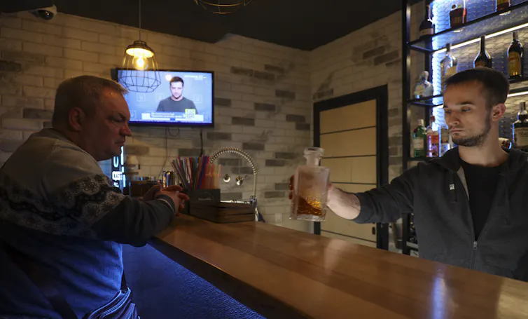Two men in an underground bar watch Ukrainian president Volodymyr Zelensky on television.
