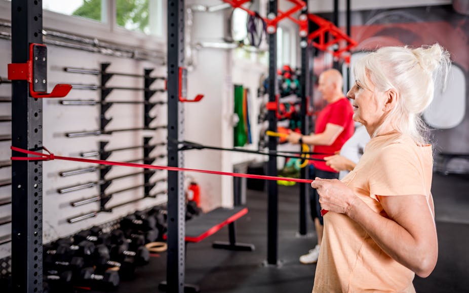 An older woman uses a rubber resistance band to train her arms while in the gym.
