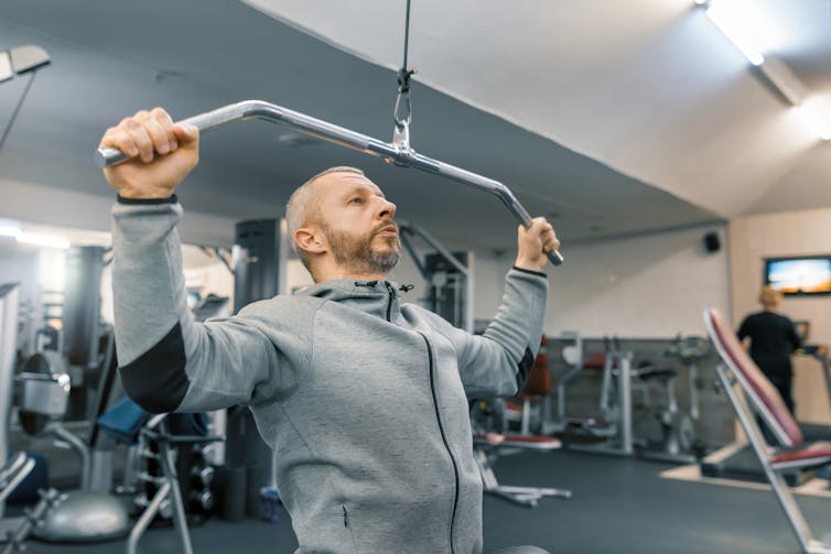 A middle-aged man performs an arm exercise at the gym using a weight machine.