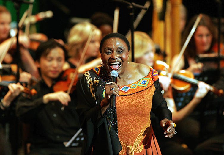 An older woman in an orange and black dress sings into a microphone, smiling, in front of a full orchestra.