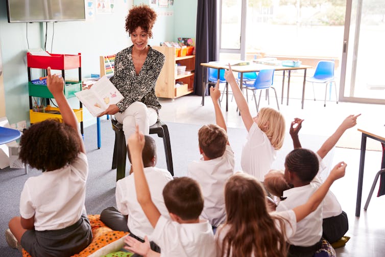 Children raising hands while teacher shows book