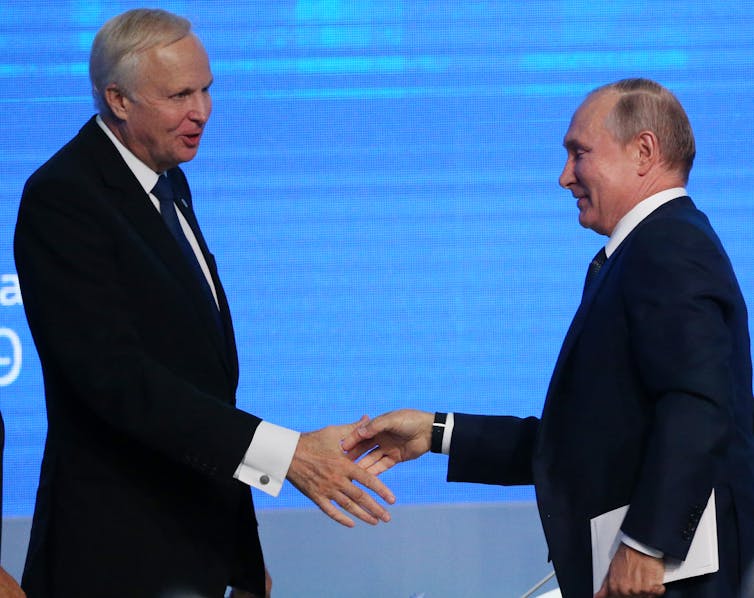 Two men in suits smile and extend hands.