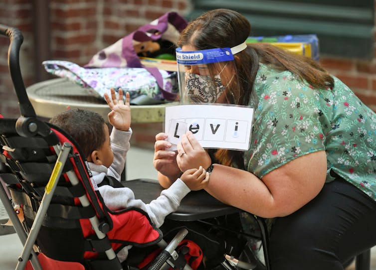 A woman wearing a face mask and a face shield holds up letters in front of a young child sitting in a stroller