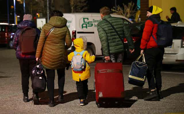 Seen from behind, a Ukrainian family walks in a line all carrying suitcases and bags and wearing winter coats