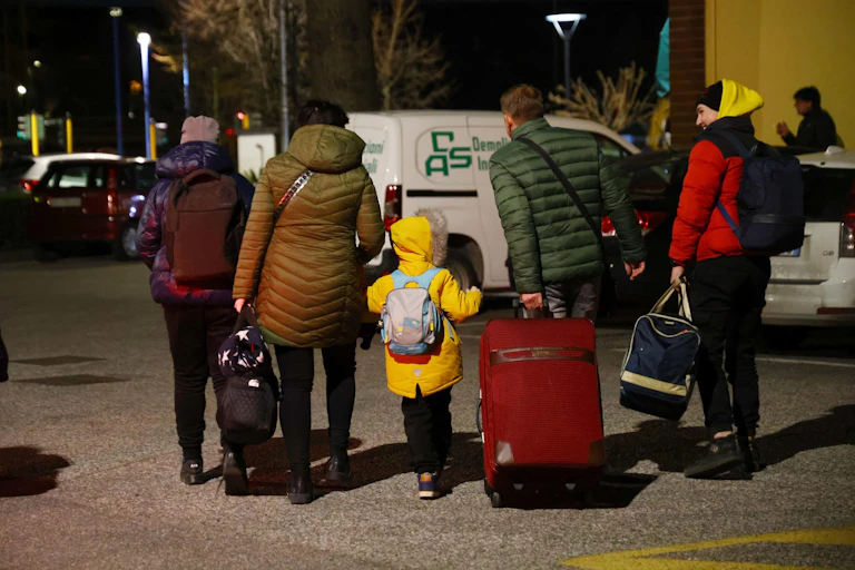 Seen from behind, a Ukrainian family walks in a line all carrying suitcases and bags and wearing winter coats