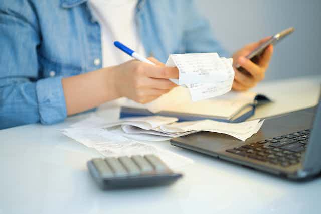 A person from the neck-down holding a receipt, pen, and mobile phone. The table in front of them has a pile of receipts, a calculator, and an open laptop on it.