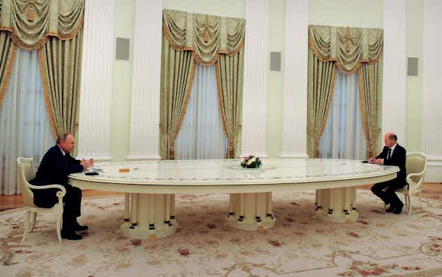 Two men sit at the opposite ends of a long table in an ornately decorated room.