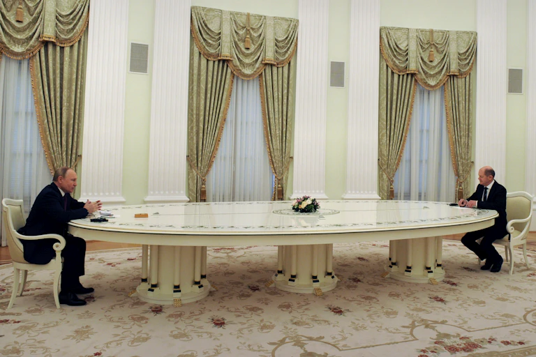 Two men sit at the opposite ends of a long table in an ornately decorated room.