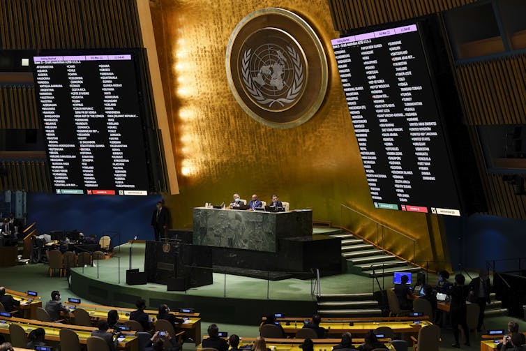 People seated in a vast hall, with a plaque bearing the United Nations logo on the wall.
