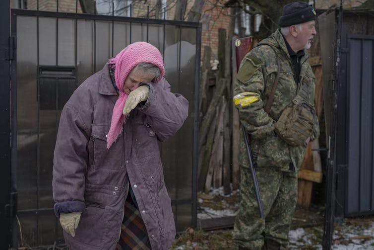 An elderly woman in a purple coat cries next to a soldier.