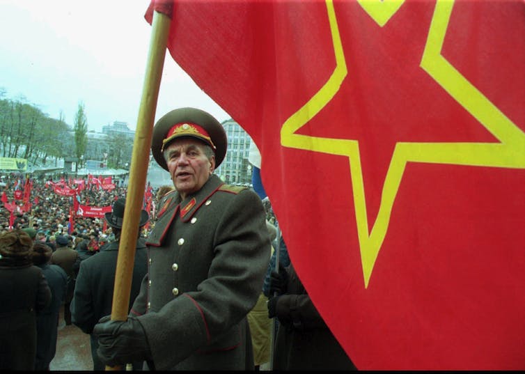 An elderly man in a military uniform holds the Soviet flag in a large gathering.