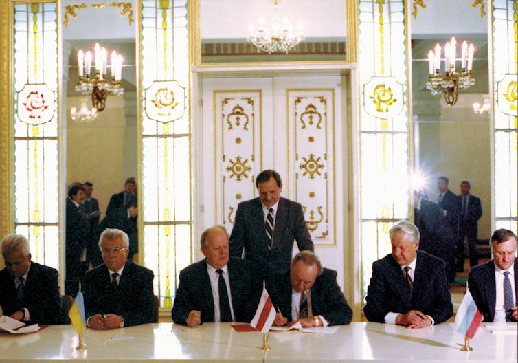 Men in suits sit at a table and sign documents in an ornate hall.