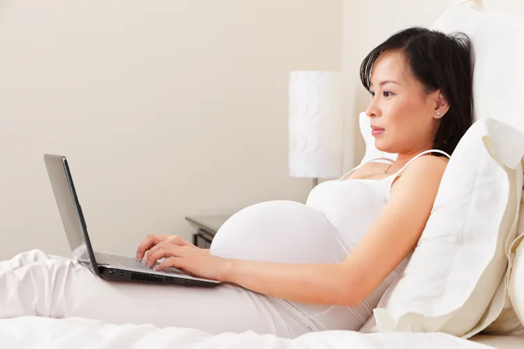 Pregnant woman wearing white, reclining on a bed while using a laptop