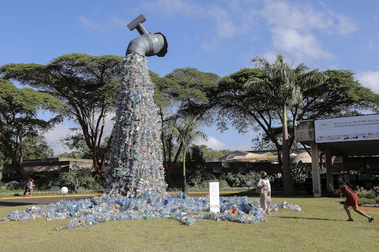 A large model of a tap pouring plastic waste onto the ground is suspended in the air before a conference centre.