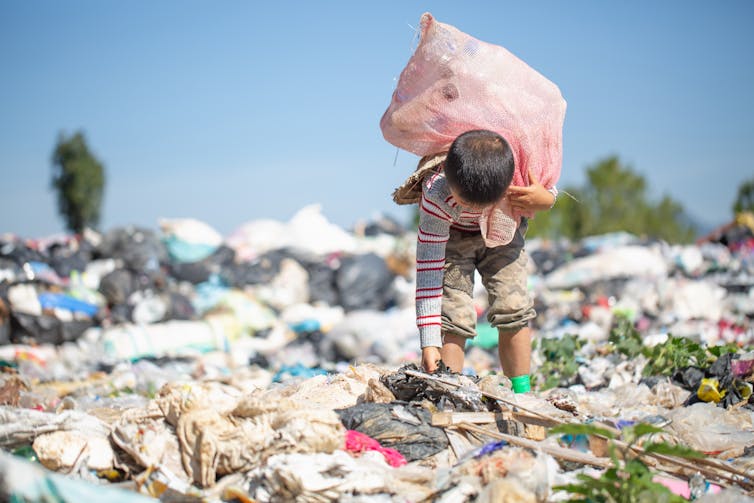 A young boy bends over to pick up litter from a plastic-strewn landscape.