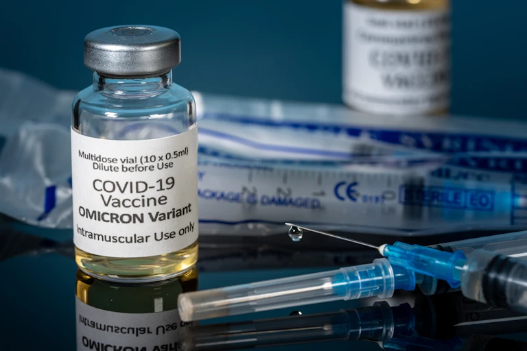 Bottle with Omicron vaccine label and syringe needle with droplet on the end.