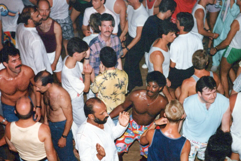 Men in a variety of 80s style clothing some with their shirts off, are seen crowded on a busy dance floor.