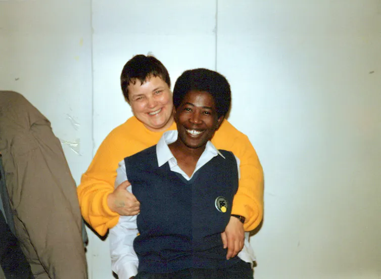 Two people smile and embrace look directly at the camera: a white butch woman wearing a bright yellow sweater wraps her arms around a black woman wearing a collared shirt and vest and lipstick.