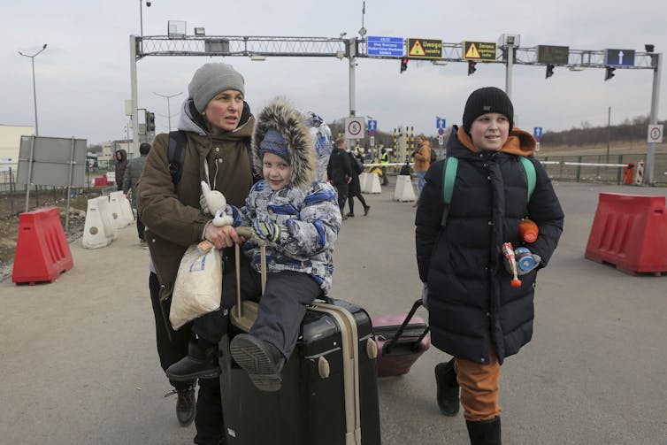 petit garçon assis sur une valise poussée par sa mère, jeune garçon qui marche à côté d’eux