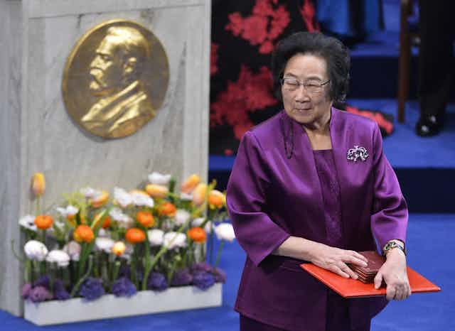 woman holds award next to podium with Nobel's profile on it