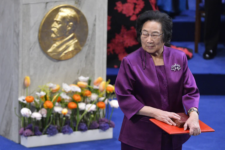 woman holds award next to podium with Nobel's profile on it