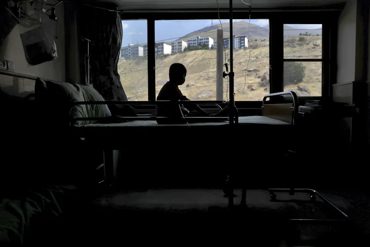 A bald boy is seen in silhouette as he looks out a window.