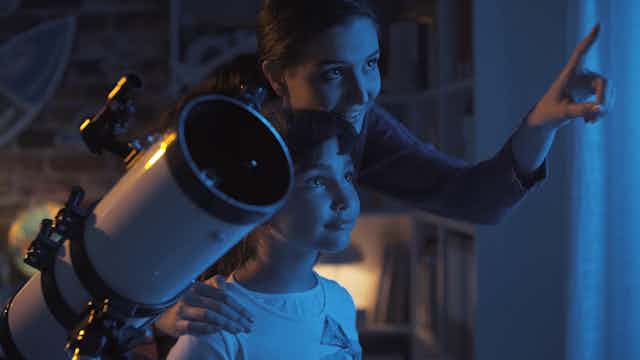 A woman and child stand in front of a window, with the woman pointing outside and upwards. A telescope is in the foreground.