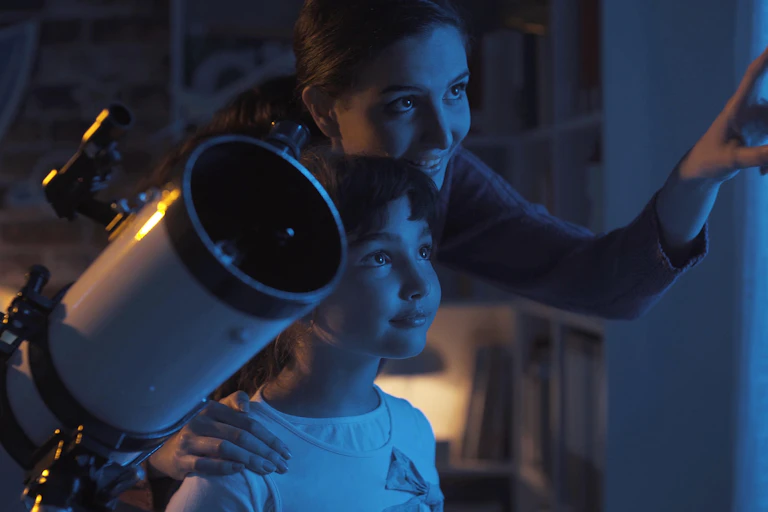 A woman and child stand in front of a window, with the woman pointing outside and upwards. A telescope is in the foreground.
