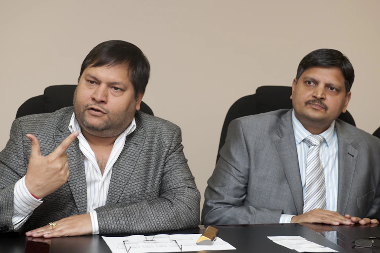 A man wearing a jacket and shirt with an open collar raises his fore and index fingers as he speaks at a press conference. Another man to his left, wearing a jacket and a tie, has both hands on a table.