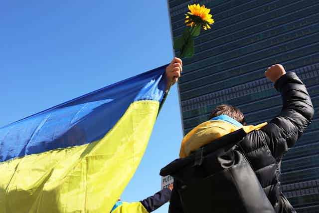 A faceless person holds a Ukrainian flag and another raises a fist outside of the UN Headquarters in NYC