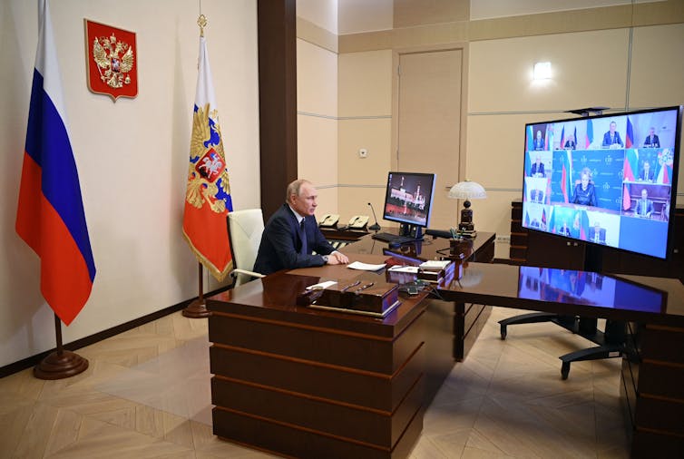 Vladimir Putin sits alone at a large wooden desk, looking at a TV screen that shows several people in a meeting.