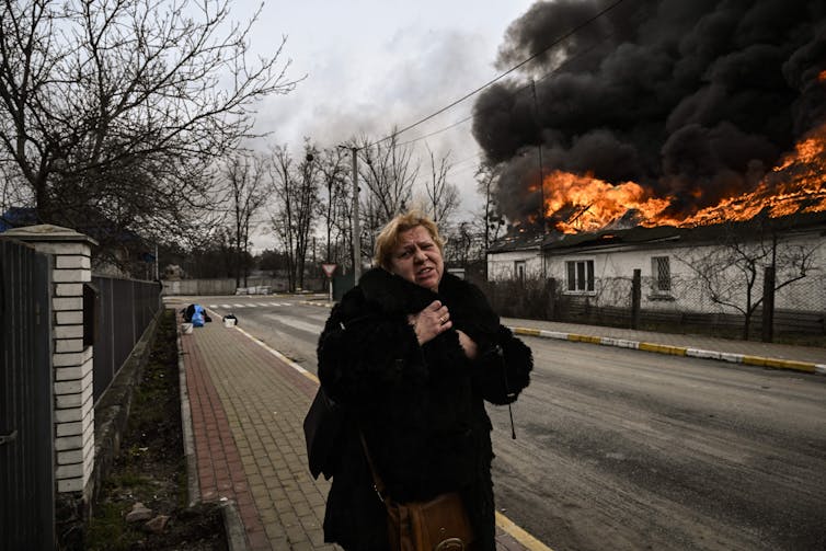 Una mujer con un abrigo oscuro llora mientras camina por una calle vacía, alejándose de un edificio en llamas.