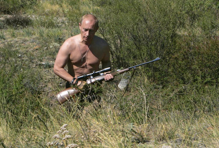 A shirtless balding man walks through a field clutching a rifle.
