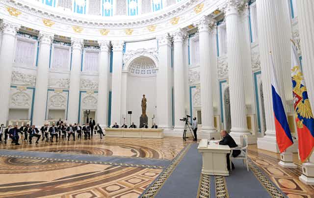 A bunch of men wearing suits sit in chairs at a significant distance from Putin, who is sitting at a white desk in a spacious room at the Kremlin 