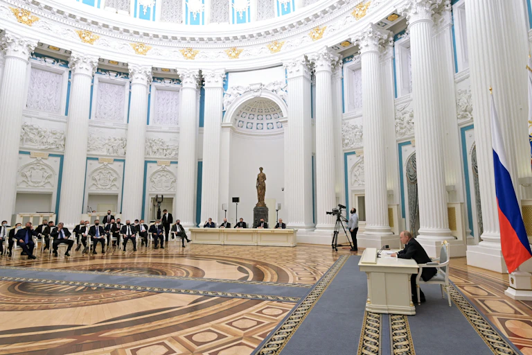 A bunch of men wearing suits sit in chairs at a significant distance from Putin, who is sitting at a white desk in a spacious room at the Kremlin