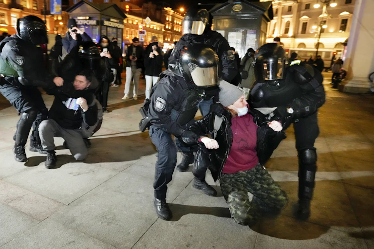 Police wearing helmets with face visors drag protesters through the street as they arrest them with St Petersburg night life in the background