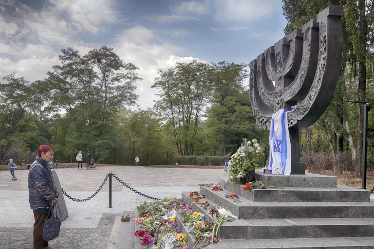 A woman stands in front of a flower-strewn Holocaust memorial featuring a large menorah on a raised platform.