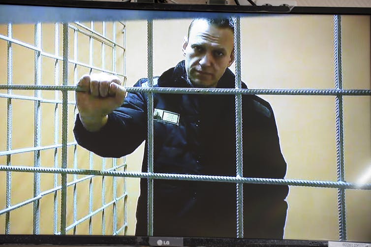 A man stands behind bars in a prison with his right hand on one of the bars