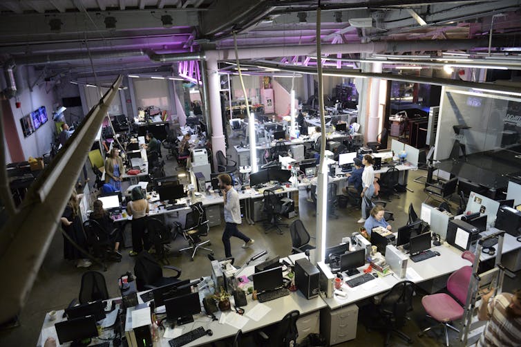 Male and female reporters work at desks in a large newsroom, some sitting, some standing, as viewed from ceiling height