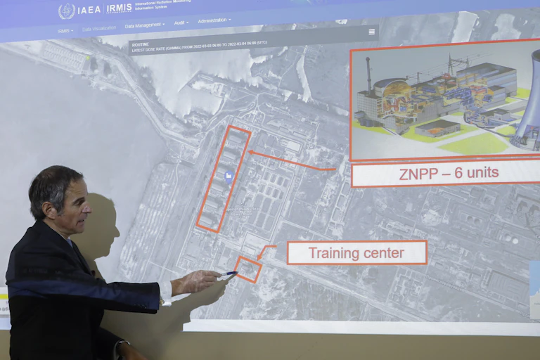 A graying man in a dark suit points to a projection on a wall of a map