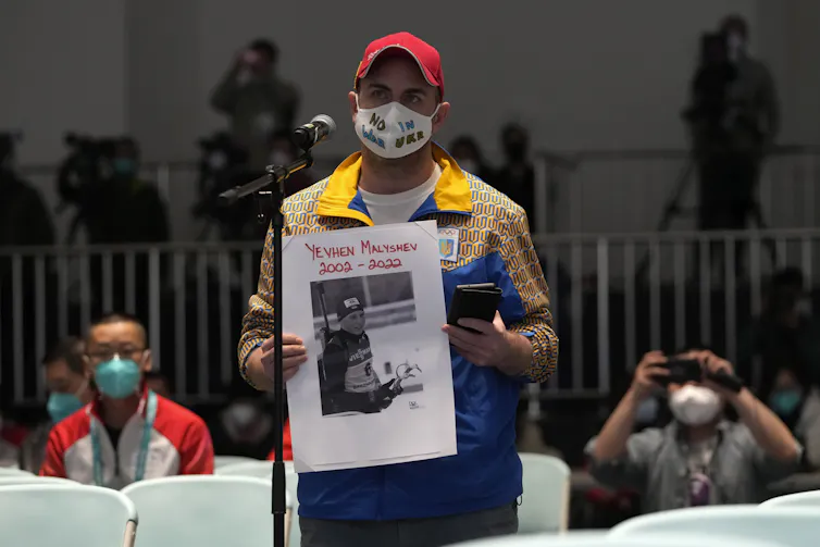 A person wearing a face mask that says 'No War in Ukraine' and holding a sign that says 'Yevhen Malyshev 2002-2022.'