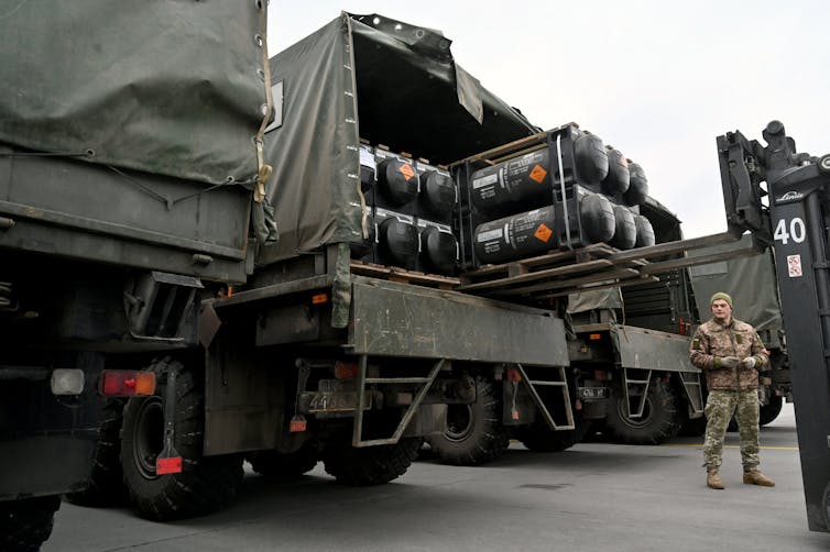 A man wearing a military uniform is standing near several large trucks that are being loaded with missiles.