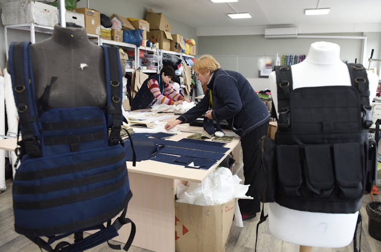 A man and a woman are sewing bullet-proof vests that are displayed near their work tables.